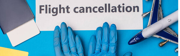 Top view of doctor in latex gloves near card with light cancellation lettering, toy plane and passport on blue background, panoramic shot