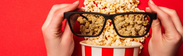 Top view of woman holding sunglasses near bucket with popcorn on red background, panoramic shot
