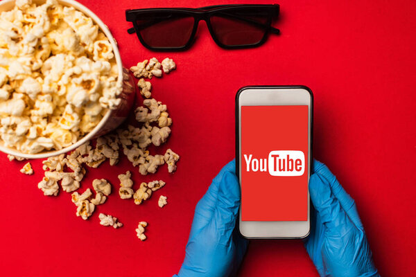 KYIV, UKRAINE - MARCH 26, 2020: Top view of person holding smartphone with youtube app near popcorn and sunglasses on red background