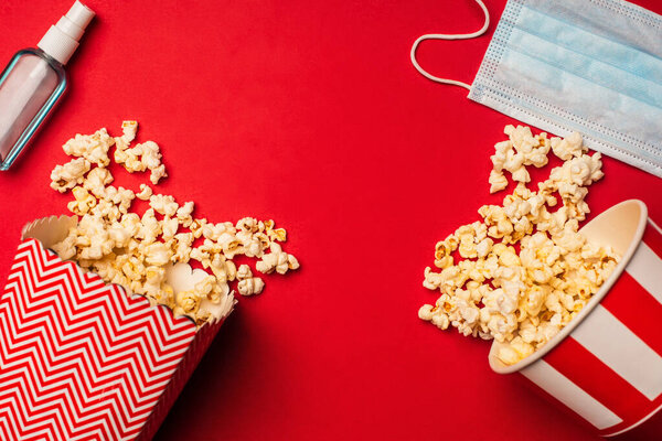 Top view of hand disanitizer, medical mask and buckets with popcorn on red surface
 