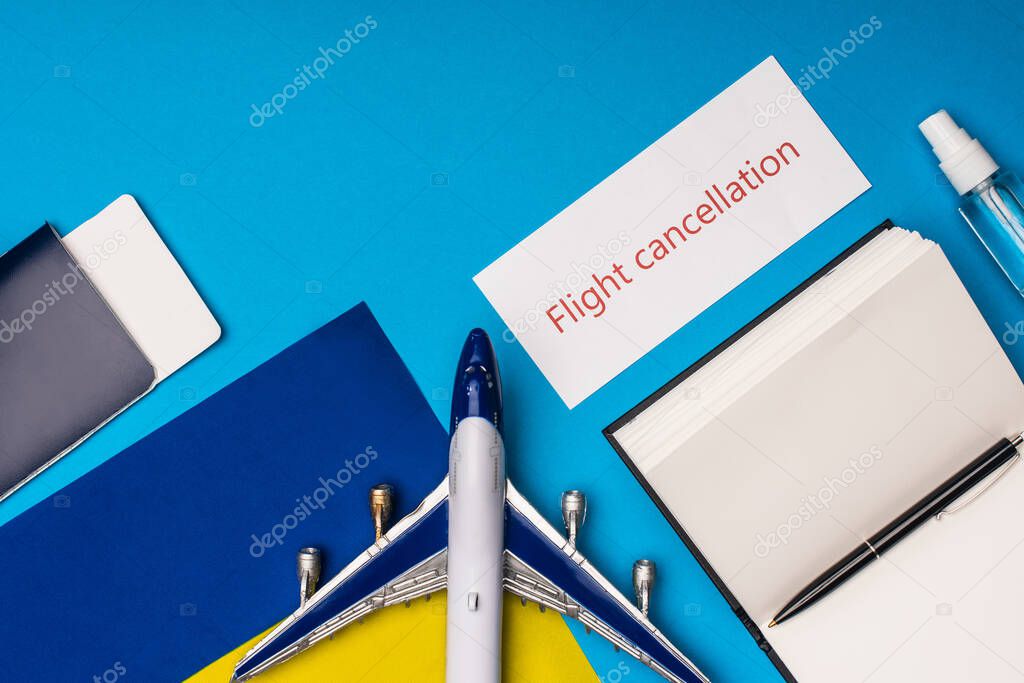 Top view of toy plane near card with flight cancellation lettering, notebook and flag of ukraine on blue background