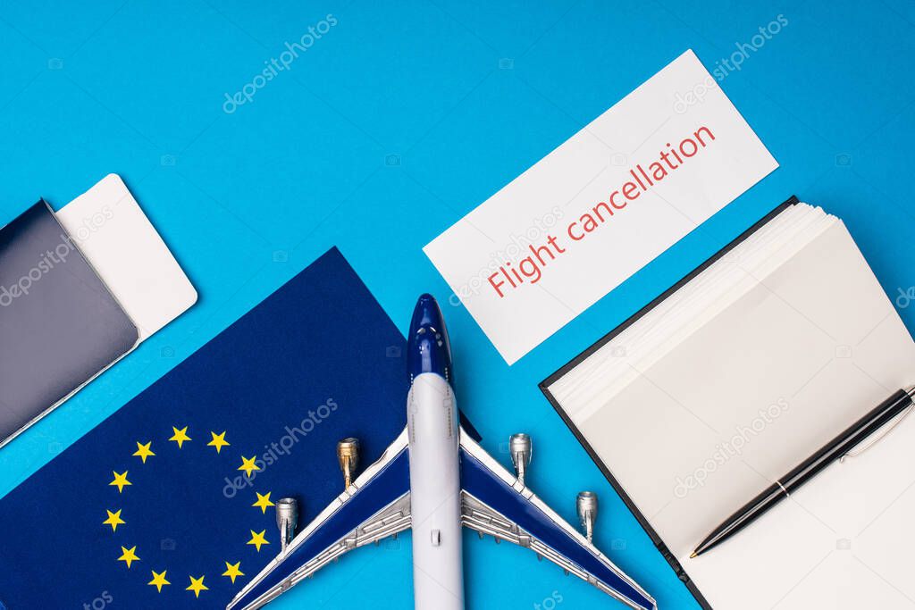 Top view of card with flight cancellation lettering near toy plane, passport and flag of european unity on blue surface