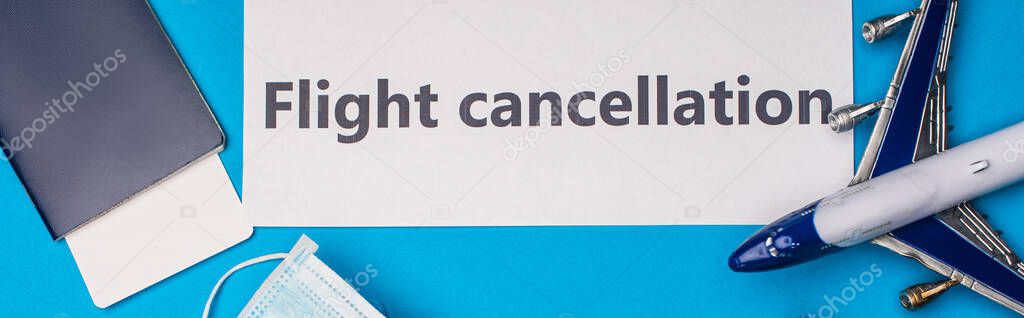 Top view of card with flight cancellation lettering near toy airplane, medical mask and passport with air ticket on blue surface, panoramic shot