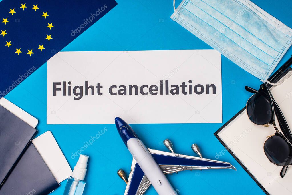 Top view of card with flight cancellation lettering near flag of european unity, toy airplane and medical mask on blue background