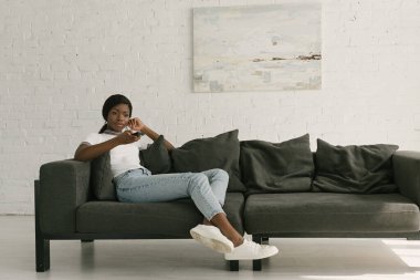 attractive african american girl holding tv remote controller while sitting on sofa at home clipart
