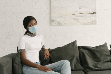 Evdeki gri kanepede oturan, tıbbi maskeli, düşünceli Afrikalı Amerikalı kız.