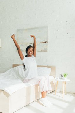 happy african american girl in pajamas stretching with raised hands while sitting on bed clipart