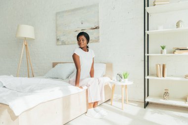 beautiful african american girl in pajamas sitting on bed and looking away in morning clipart