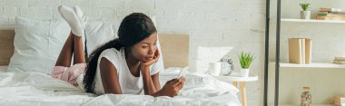 Afro-Amerikan kız panoramik ürünleri yatakta uzanıyor ve akıllı telefondan sohbet ediyor.