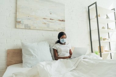 Tıbbi maskeli Afrikalı Amerikalı serbest yazar dizüstü bilgisayarla yatakta oturup kameraya bakıyor.