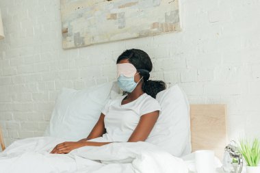 Tıbbi maskeli ve uyku maskeli genç Afrikalı Amerikalı kadın yatakta dinleniyor.