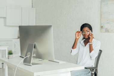 Ciddi bir Afro-Amerikan serbest çalışanı akıllı telefondan evde bilgisayar monitörü başında konuşuyor.