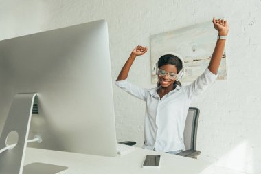 Mutlu Afrikalı Amerikalı serbest çalışan, bilgisayar monitörünün yanında kablosuz kulaklıkla müzik dinlerken ellerini kaldırarak el kol hareketi yapıyor. 