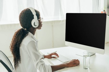 Genç Afrikalı Amerikalı kablosuz kulaklıklı serbest çalışan boş ekranlı bilgisayar monitörüne bakıyor.