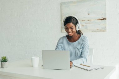 Gülümseyen Afrikalı Amerikalı kablosuz kulaklıklı, evde dizüstü bilgisayarla çalışan.