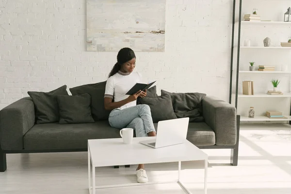 ノートパソコンとテーブルの近くのソファに座っている間にアフリカ系アメリカ人のフリーランサーがノートを読む — ストック写真