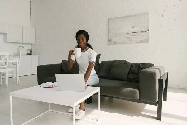 幸せなアフリカ系アメリカ人のフリーランスがカメラで笑顔でソファに座りながらノートパソコンとテーブルの近くにコーヒーを飲みながら — ストック写真