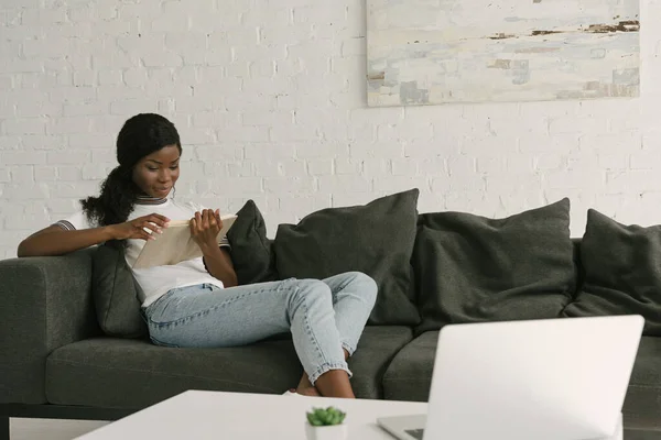 Sonriente Afroamericano Freelancer Sentado Sofá Leer Libro Cerca Mesa Con — Foto de Stock