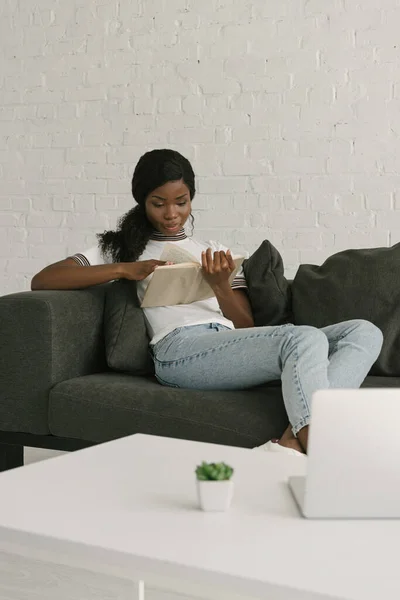 Joven Afroamericano Freelancer Descansando Sofá Mientras Lee Libro Cerca Mesa — Foto de Stock