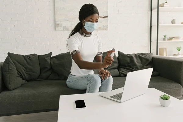 医療用マスクの若いアフリカ系アメリカ人のフリーランスがノートパソコンに防腐剤を散布し — ストック写真