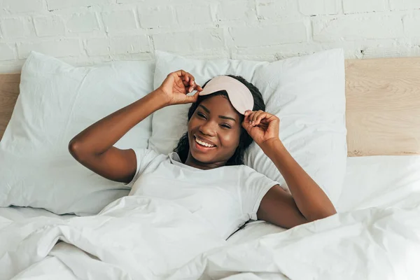Uśmiechnięta Afroamerykanka Dotykająca Maski Snu Leżąc Łóżku Patrząc Kamerę — Zdjęcie stockowe