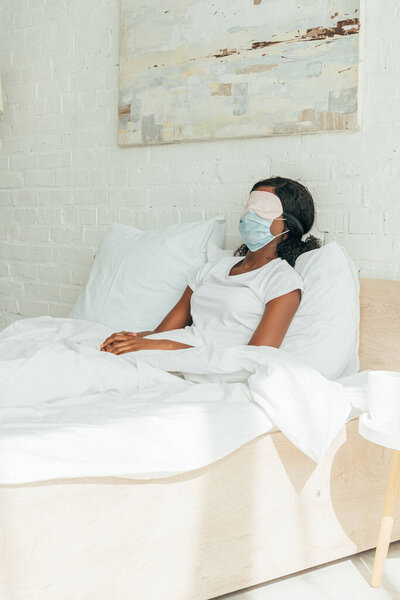 african american girl in medical mask and sleep mask resting while sitting in bed