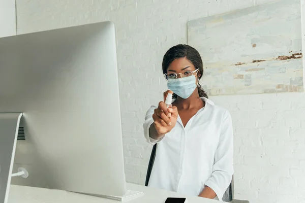 医療用マスクを着た若いアフリカ系アメリカ人のフリーランスは 自宅のコンピュータモニタの近くに座っている間に防腐スプレーを示す — ストック写真