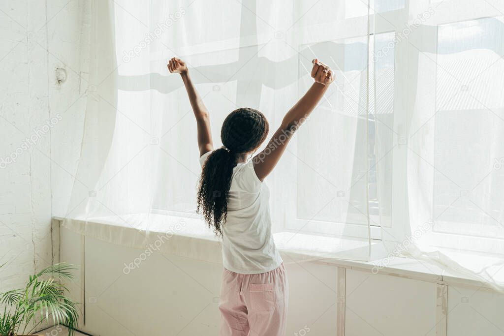 back view of african american girl in pajamas stretching hands while standing near window in sunlight
