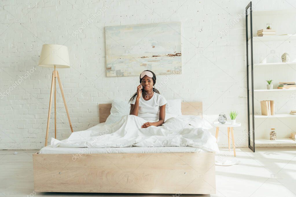 young african american woman with sleep mask on forehead sitting in bed while talking on smartphone