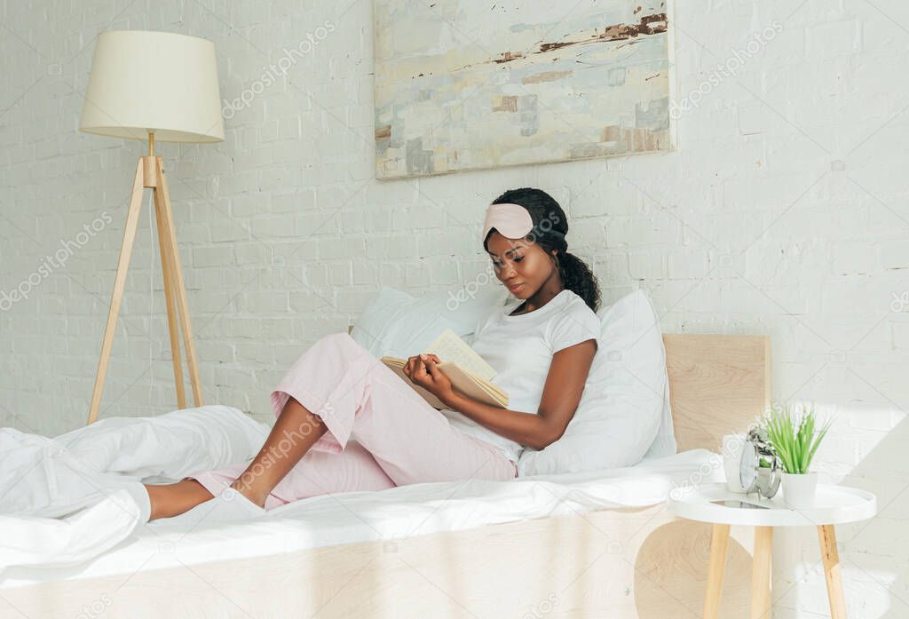 attentive african american girl with sleep mask on forehead reading book while sitting in bed