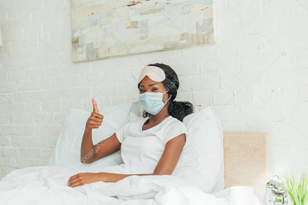 african american girl in medical mask showing thumb up while looking at camera 