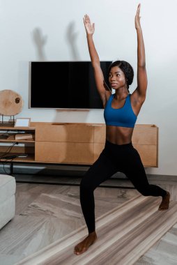 Afrika kökenli Amerikalı sporcu kadın yüksek hamlede duruyor elleri havada ve oturma odasında yoga minderinde gülümsüyor.