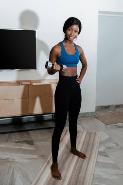 African american sportswoman with hand on hip training with dumbbell on fitness mat, smiling and looking at camera in living room clipart