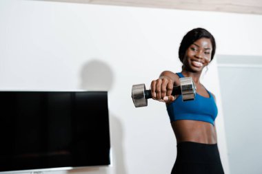Afrika kökenli Amerikalı sporcu kadın, uzanmış elleriyle dambılı tutuyor, gülümsüyor ve oturma odasında kameraya bakıyor.