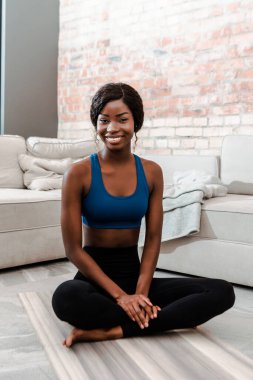 African american sportswoman in lotus pose smiling and looking at camera on yoga mat in living room clipart