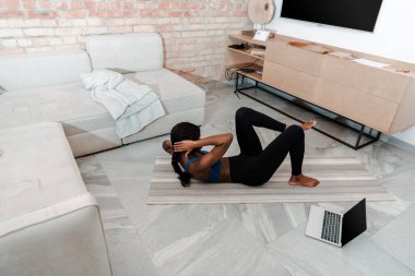 Afrika kökenli Amerikalı sporcunun oturma odasındaki laptopun yanında yoga minderinde karın kasları yaparken yüksek açılı görüntüsü.