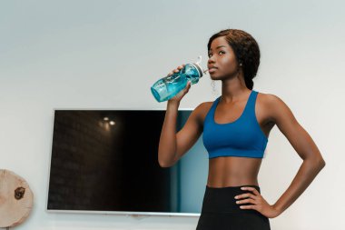 Oturma odasında eli kalçasında su içen Afrikalı Amerikalı sporcu kadın.