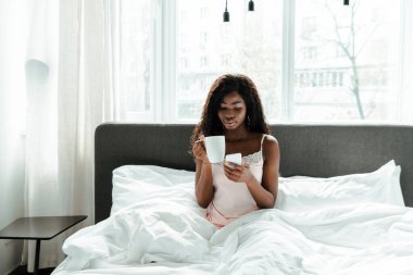 Afrika kökenli Amerikalı kadın yatak odasında elinde çay ve akıllı telefon tutuyor.