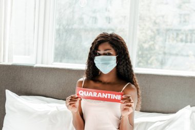 Tıbbi maskeli Afrikalı Amerikalı bir kız yatak odasında karantina kağıtları olduğunu gösteriyor.