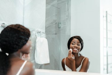 African american woman wiping face with cotton pad, looking at mirror and smiling in bathroom clipart