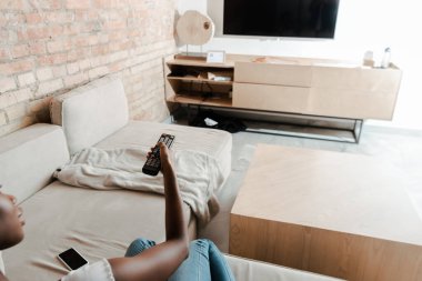 Uzaktan kumandayı elinde tutan ve oturma odasında televizyonu gösteren Afro-Amerikan kadın görüntüsü.