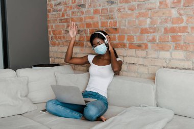 Oturma odasındaki kanepede elleri, kulaklıkları ve laptopuyla tıbbi maskeli Afrikalı Amerikalı serbest çalışan.