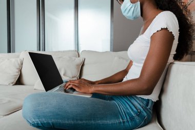 Tıbbi maskeli Afro-Amerikalı 'nın oturma odasındaki kanepede dizüstü bilgisayarla çalışmasını izledik. 