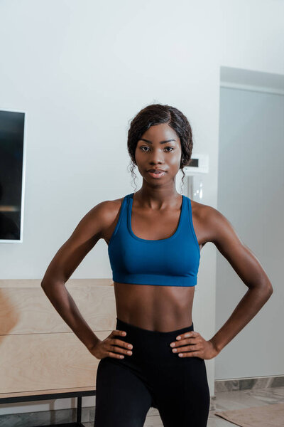 Front view of african american sportswoman with hands on hips doing lunges and looking at camera in living room