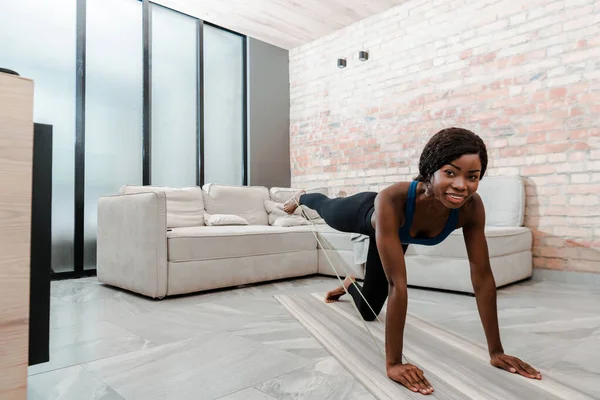 African american sportswoman on all fours practicing yoga with resistance band, smiling and looking at camera in living room