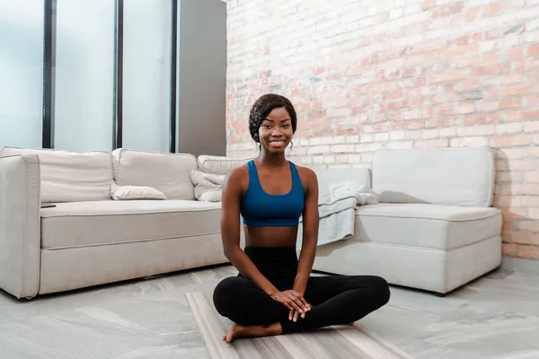 在客厅里 非洲裔美国女运动员面带微笑 看着镜头 坐着荷花在瑜伽垫上摆姿势 — 图库照片