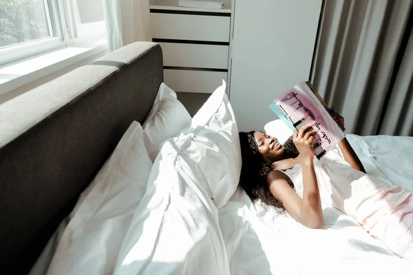 Vista Ángulo Alto Mujer Afroamericana Leyendo Revista Sonriendo Cama Dormitorio —  Fotos de Stock