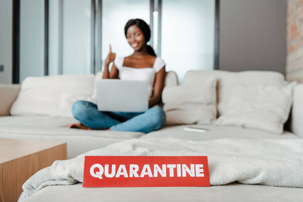 Избирательный фокус африканского фрилансера с ноутбуком, показывающим как знак и улыбающимся на диване с карточкой с карантинными помещениями в гостиной