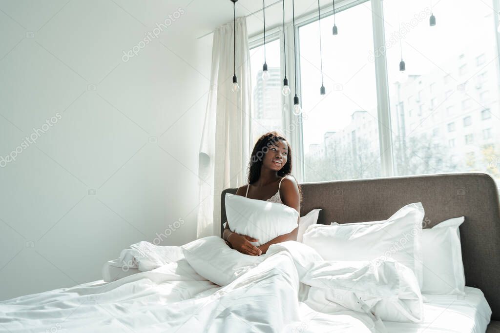 African american woman holding pillow and looking away on bed in bedroom