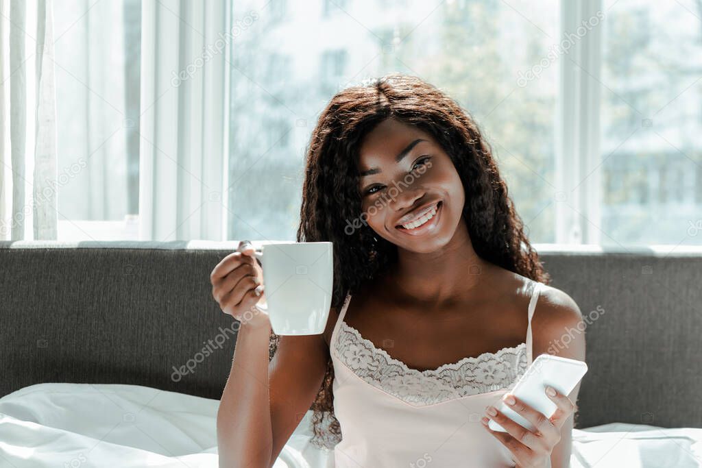 African american woman with smartphone smiling, showing cup of tea and looking at camera on bed in bedroom